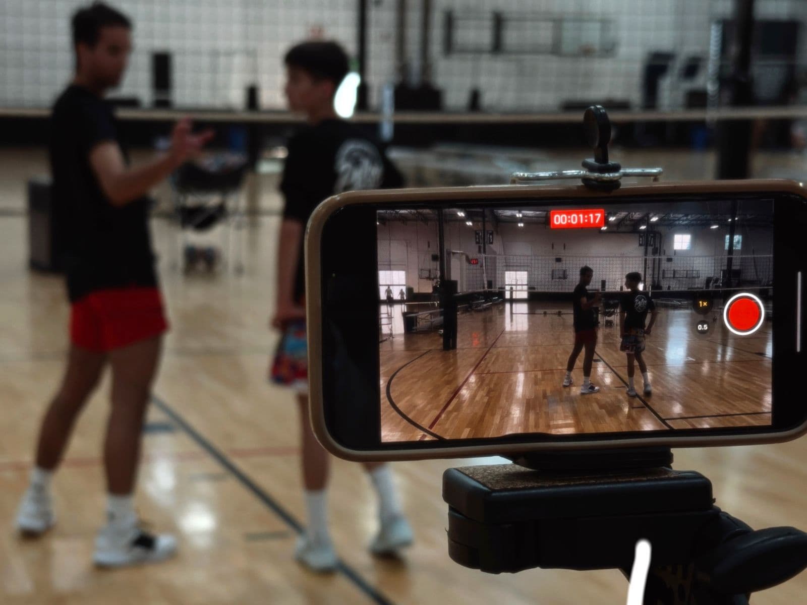 A coach talking an athlete through a drill on an indoor court while a phone on a tripod records the rep